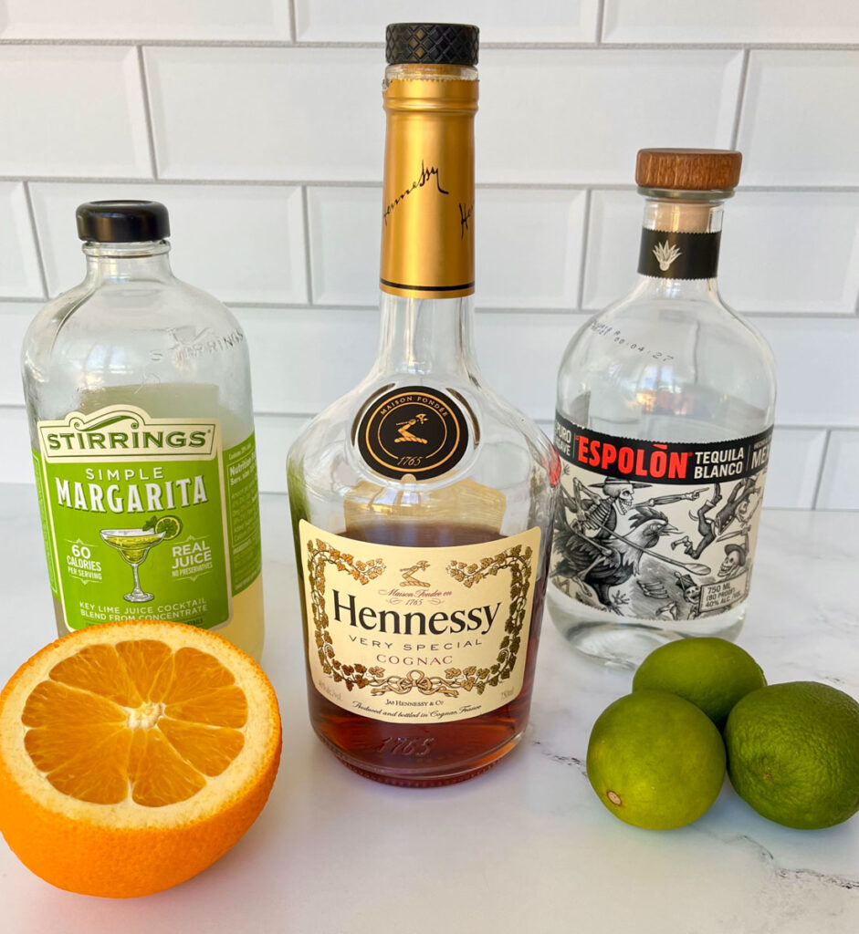 There is a bottle of Hennessy Cognac. There is a bottle of margarita mix to the left of the Hennessy bottle. There is a bottle of tequila blanco to the right of the Hennessy bottle. There is a half of an orange and 3 limes lying in front of the bottles. Items are on a white marble surface with a white tile back splash.