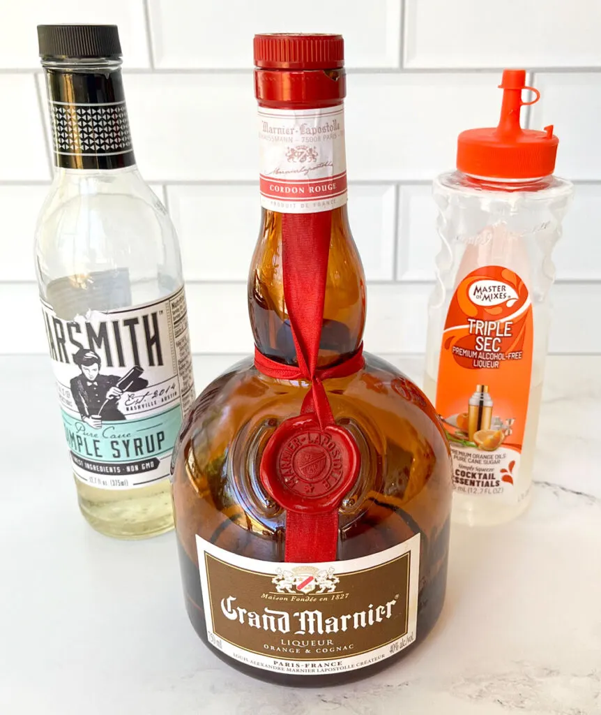 Bottle of Grand Marnier with a bottle of simple syrup to the left it and a bottle of triple sec to the right of it. Bottles are on a white marble surface in front of a white tile backsplash.