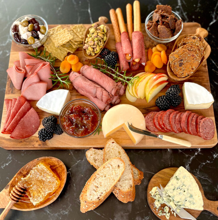 There is a large rectangular wood board with various food items on it. Meats: rolled slices of roast beef, sliced beef sausage, thin slices of folded in half beef salami and slices of beef bologna folded in triangles. Cheese: There are 2 wedges of brie cheese, a wheel of smoked gouda on the board. There is a wedge of blue cheese on a small round wood board in front of the large rectangular board. There is a small wood bowl with pistachio nuts, another wood bowl with brown crackers in it. There are 3 small clear glass bowls on the board. One with brown colored fruit spread, one with olives and chunks of feta cheese and the third bowl has pieces of beef jerky in it. There are blackberries, sliced apples, and orange colored dried apricots. There is a wood bowl with a honey comb and honey dipper sitting in front of the board. There are three slices of crusty bread lying in front of the board. The board and items are on a black marble surface.