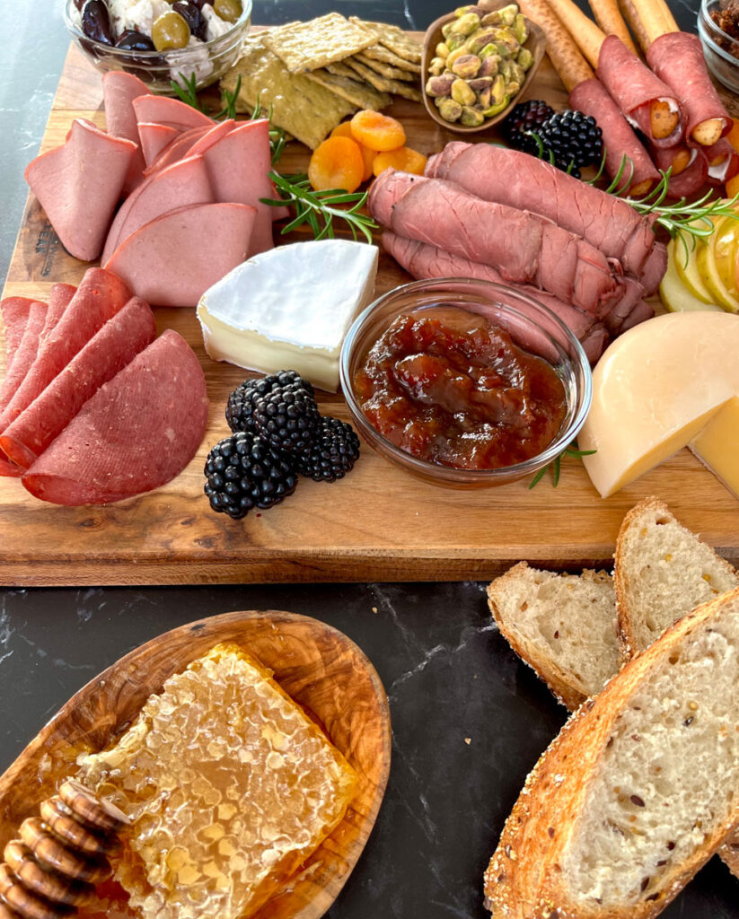 There is a wood board. On the wood board there is sliced of roast beef rolled up, slices of beef sausage. a wheel of light yellow gouda cheese, apple slices, blackberries, and dried apricots, a pile of light brown crackers, a small clear glass bowl with green olives and chunks of white feta cheese, a wood bowl with pistachios, thin bread sticks with dried beef wrapped around them. There are 3 slices of rustic bread and a wedge of blue cheese in front of the board and a wood bowl with a honey comb in it. Items are on a black marble surface.