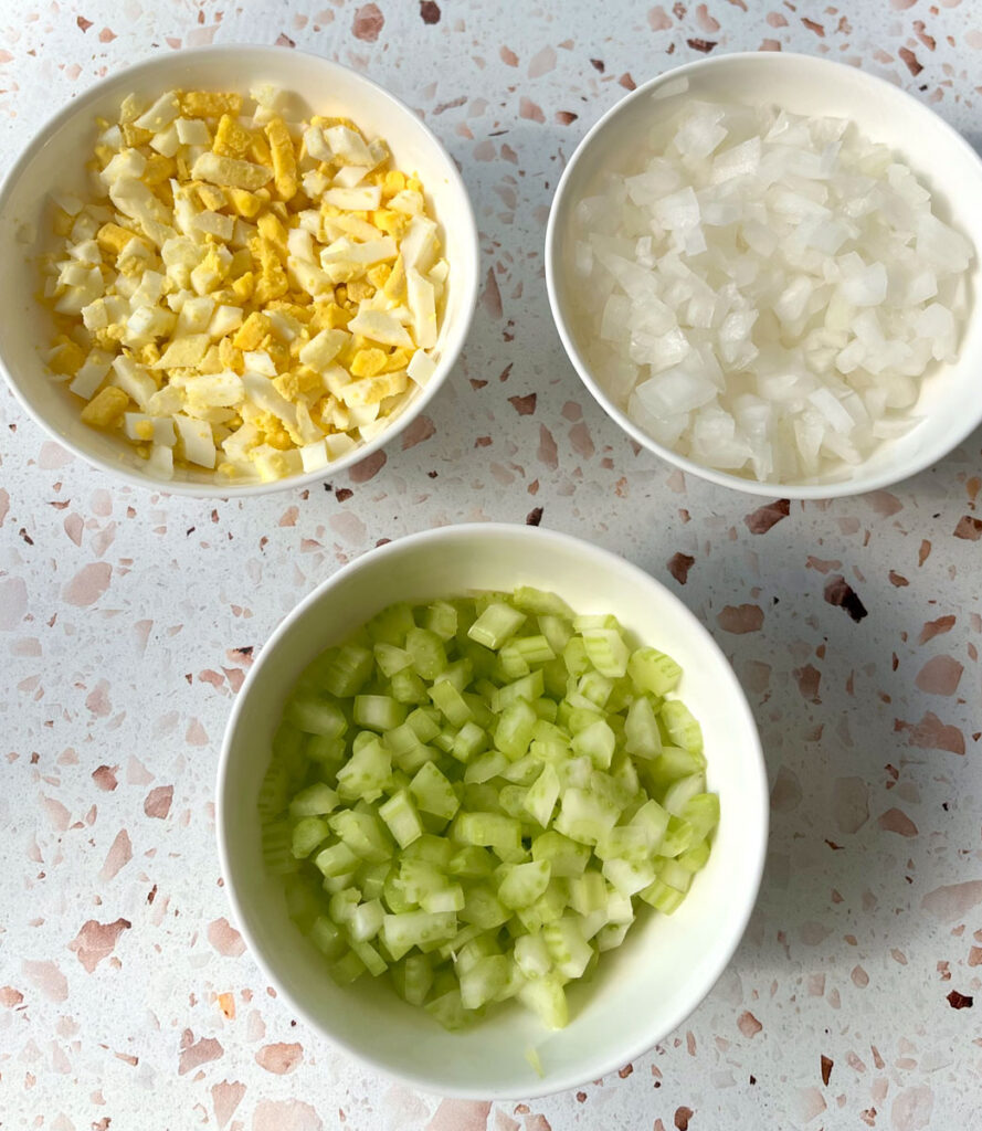 There are 3 exact same size white bowls. One has chopped celery, one has chopped white onion, and the third one has chopped hard boiled egg in it. Bowls are on a light red terrazzo surface.