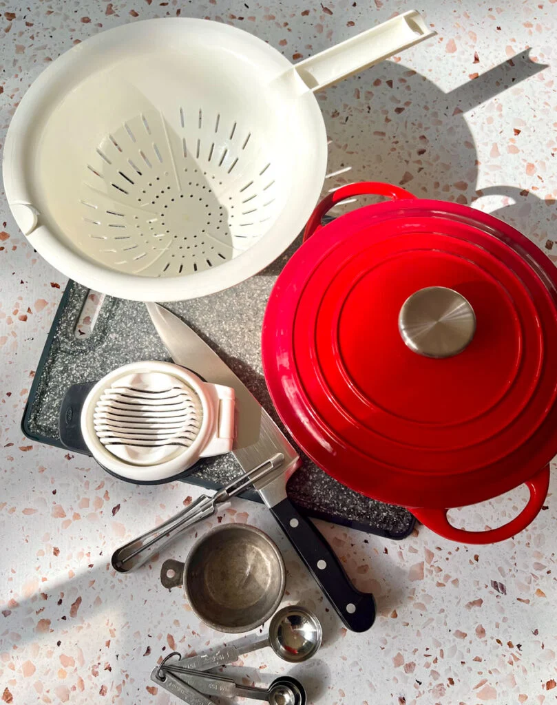 There is a gray cutting board, a white plastic colander, a large red pot with a lid, an egg slicer, vegetable peeler, large kitchen knife, a measuring cup, and a set of measuring spoons. All items are on a light red terrazzo surface.