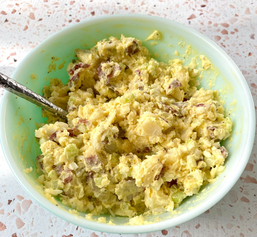 White mixing bowl with chopped red skinned potatoes, chopped celery, chopped hard boiled eggs, and chopped onion, mayonnaise, mustard, salt, and pepper mixed together in it. There is a silver spoon resting in the bowl. The bowl is on a light red terrazzo surface.