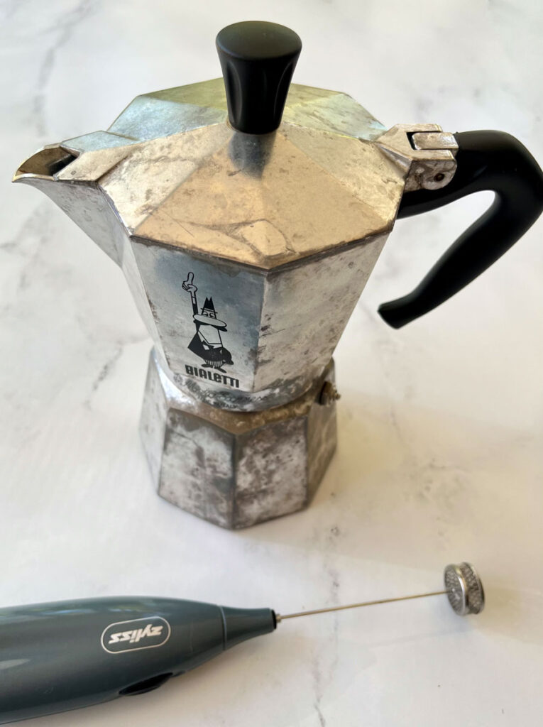 There is a stainless steel moka coffee pot and a gray handheld mild frother laying in front of the pot. Items are on a white marble surface.