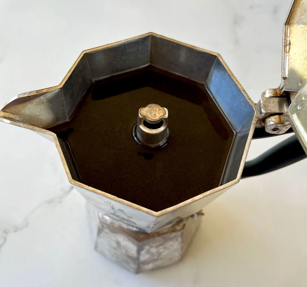 There is a stainless steel moka coffee pot with the lid open. There is brewed coffee in the top chamber. Pot is on a white marble surface.