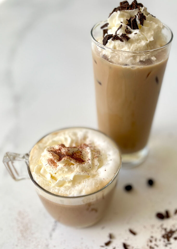 There is a clear cup with a light brown coffee milk mixture in it. There is white milk foam on top of the liquid mixture as well as whipped cream and sprinkled cocoa powder. There is a tall clear glass behind the cup with the same coffee and milk mixture in it. There is whipped cream and dark chocolate bits on top of the coffee. Items are on a white marble surface. There is coffee beans and chocolate bits scattered on the surface in front of cup and glass.