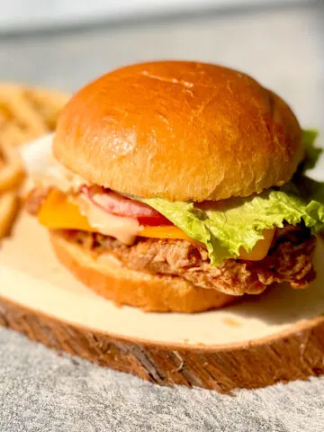 There is an oval piece of wood with a chicken sandwich on it. The chicken sandwich is a round bun with a piece of fried chicken, a slice of cheddar cheese, a slice of tomato, a piece of lettuce and light orange sauce on it. There are french fries on the piece of wood. The piece of wood is on a light gray surface.