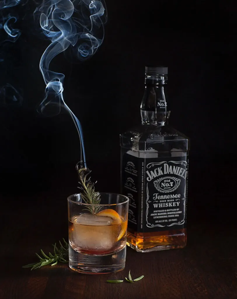 There is a large bottle of Jack Daniels Whiskey. Next to the bottle is a short clear glass with brown liquor, a large ice cube, an orange peel and a green sprig of rosemary in it. The sprig of rosemary has smoke coming off the top. There are pieces of rosemary laying around the glass. Items are on a black surface.