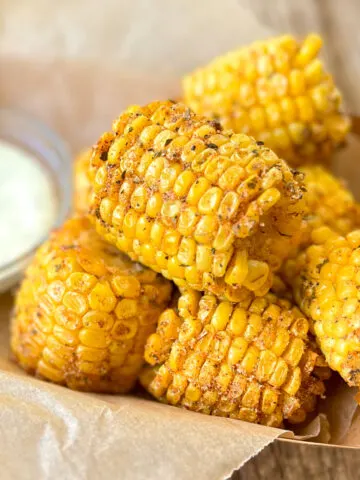 Six small pieces of fried corn on the cob with light brown seasoning on them. The corn is in a light brown food box lined with a light brown piece of paper. There is a small clear bowl with white sauce sitting next to the pieces of corn in the food box. The food box is on a wood surface.
