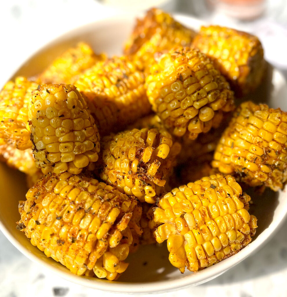 There are small pieces of fried corn on the cob with light brown dry seasoning on them. The pieces of corn are in a white serving bowl. Bowl is on a black and white terrazzo surface.