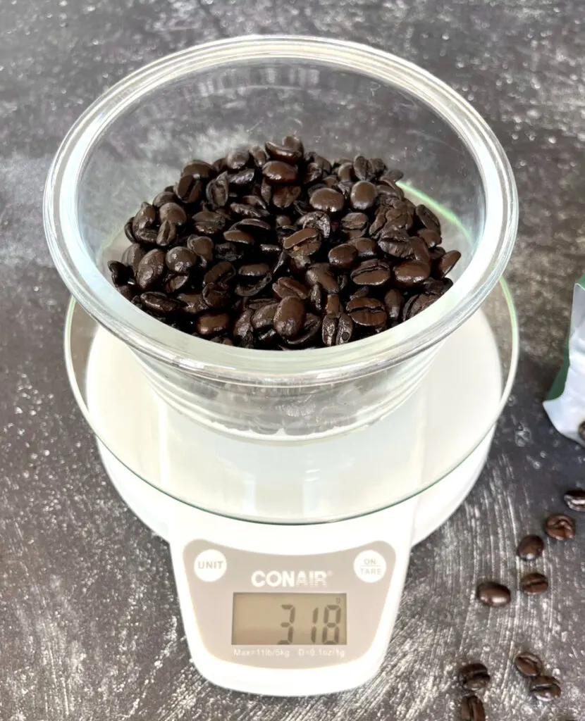 There is a white round digital kitchen scale with a clear glass bowl on it. There are coffee beans in the clear glass. There are scattered coffee beans lying next to the scale. Items are on a dark gray surface.