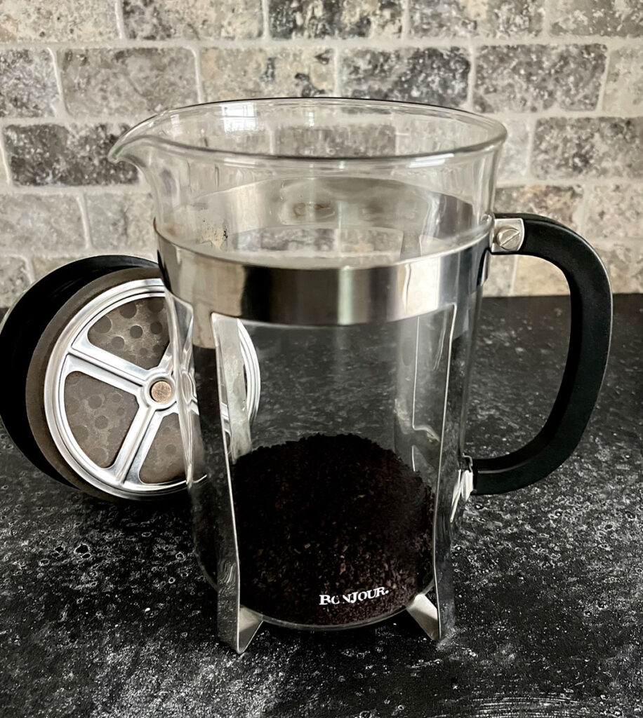 There is a clear glass cylindrical carafe with a stainless steal frame holder. There is ground coffee in the bottom of the carafe. The plunger top for the carafe is lying next to the carafe. Items are on a dark gray surface with a tile back wall.
