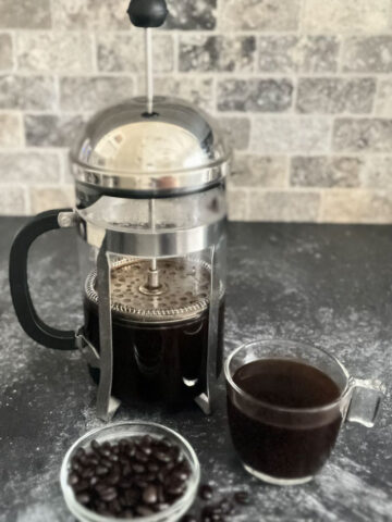 There is a clear glass cylindrical with a stainless steel plunger top with dark liquid coffee in it. There is a clear coffee cup with dark liquid coffee in it in front of the glass cylinder. There is a small clear glass bowl with coffee beans in it. All items are on a dark gray surface with a tile back wall.