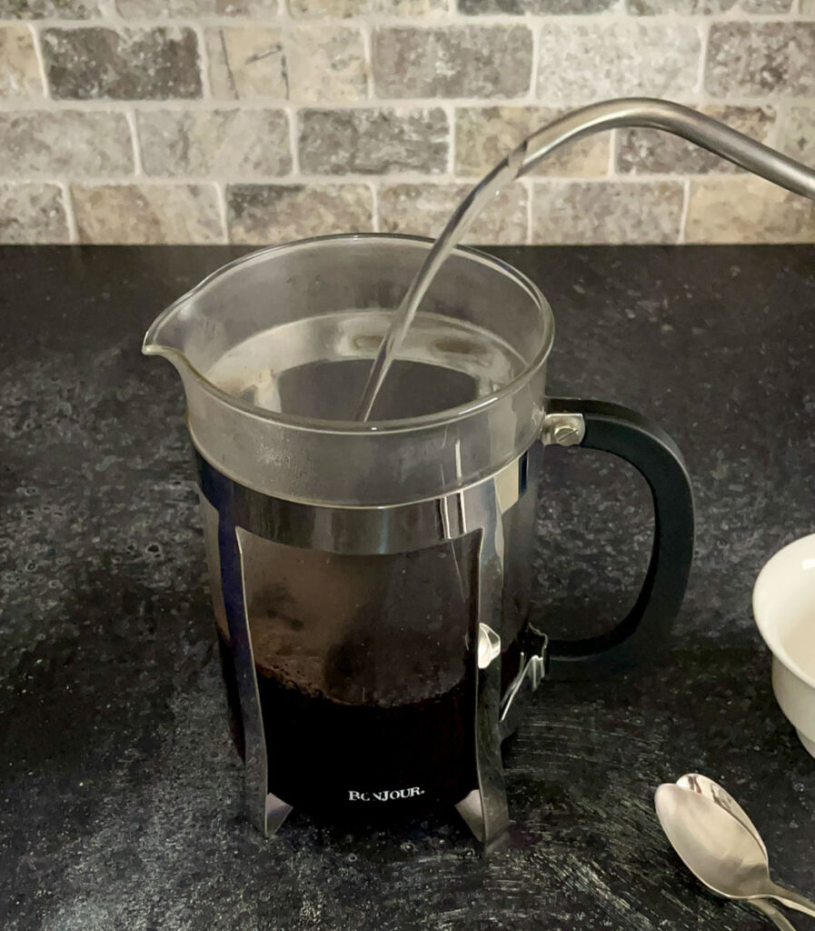There is a clear glass cylindrical carafe with a stainless steal frame holder. There is a spout with water pouring out over the carafe and into the carafe. There are 2 stainless steel spoons lying next to the carafe. The items are on a dark gray surface with a tile back wall.