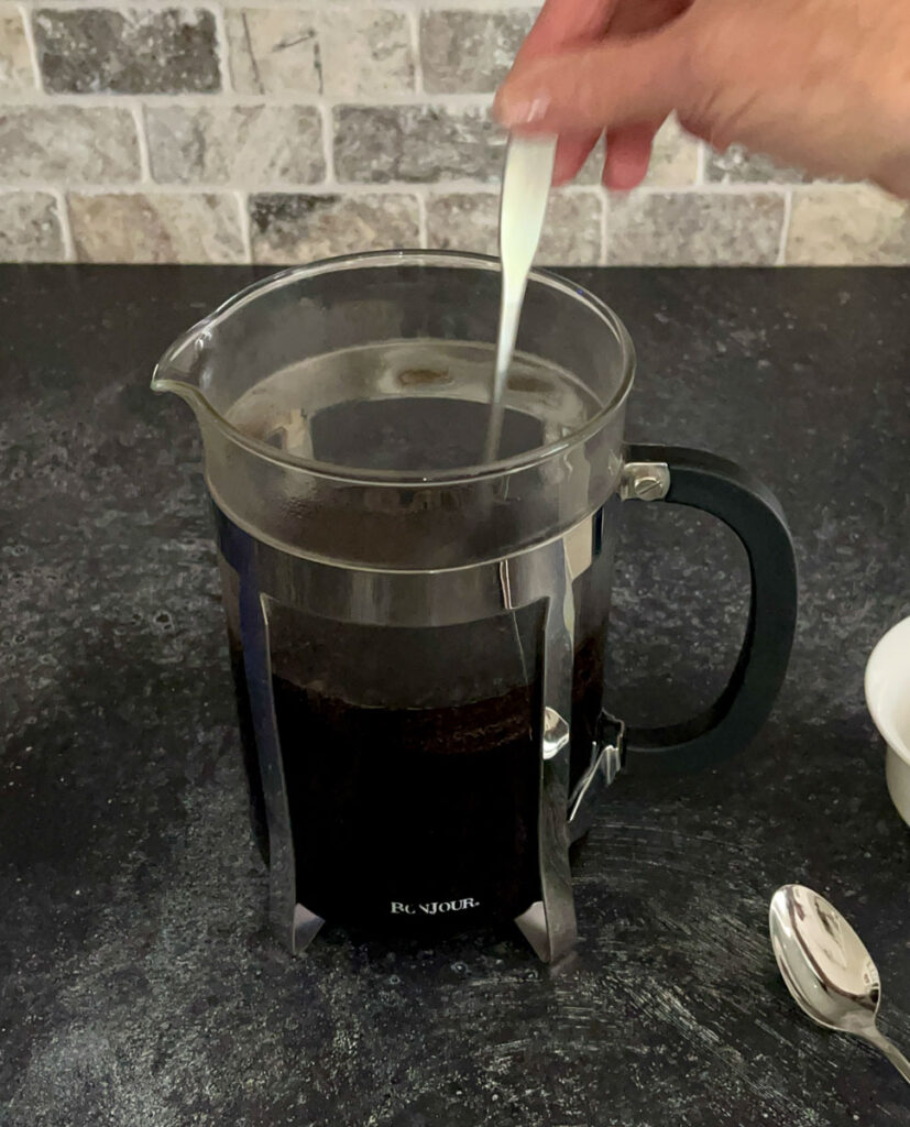 There is a clear glass cylindrical carafe with a stainless steal frame holder. There is a hand stirring the coffee with a spoon. There is a spoon lying next to the carafe. The items are on a dark gray surface with a tile back wall.