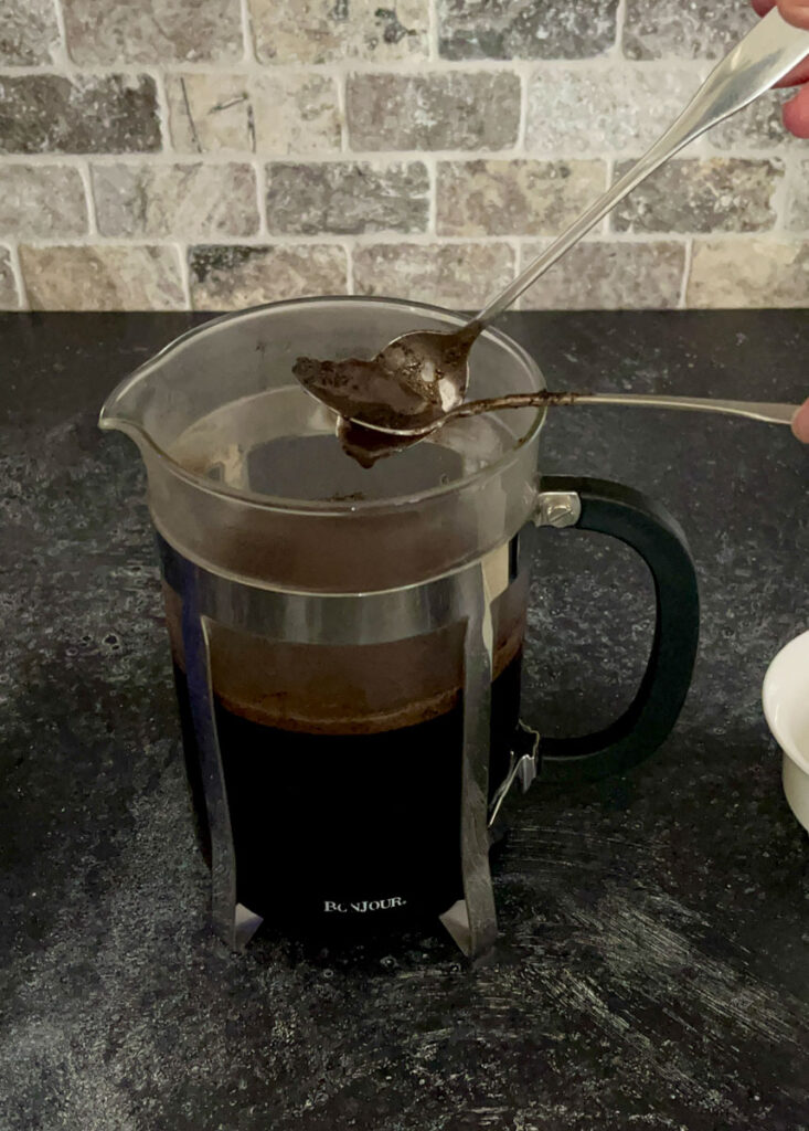 There is a clear glass cylindrical carafe with a stainless steal frame holder. There is a hand using 2 spoons to remove coffee grounds form the cylinder. The carafe is on a dark gray surface with a tile back wall.