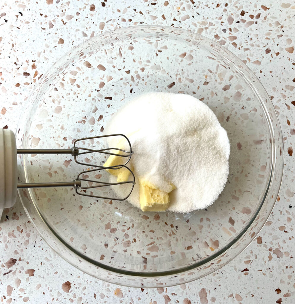 Large clear glass mixing bowl with two sticks of butter and white sugar in it. There are mixer beaters resting in the bowl. Items are on a red and white terrazzo surface.