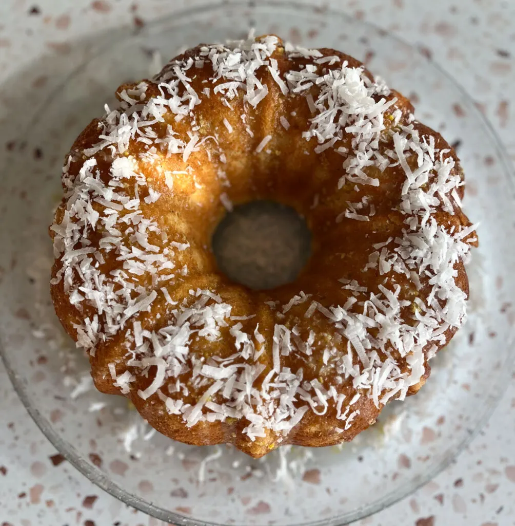 There is a clear glass round plate with a light brown doughnut shaped cake with a light glaze and shredded coconut on top of it. Plate is on a red and white terrazzo surface.