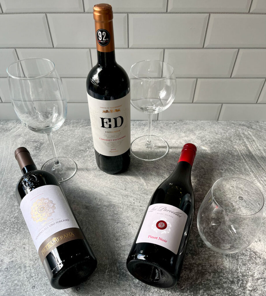 There are three bottles of red wine. Two of the wine bottles are laying down. The third wine bottle is standing upright in between them. There are two stemmed wine glasses and one without a stem. The glasses are standing next to the wine bottles. Items are on a light gray surface with a white tile back wall.