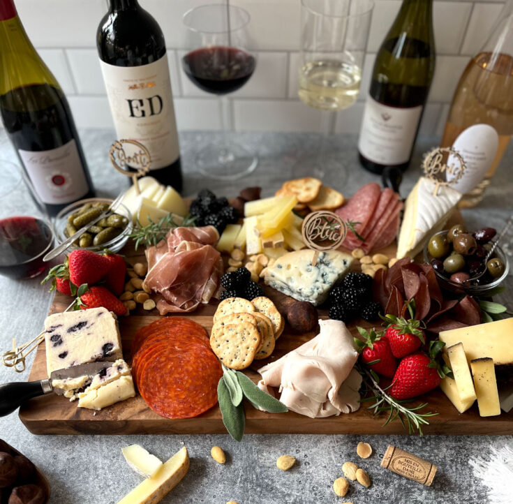 There is a large wooden board with a variety of food items: Meats, cheeses, strawberries, blackberries, nuts, olives, crackers, chocolate truffles, and fresh green herbs. There are four bottles of wine and three clear wine glasses. Items are on a light grey surface with a white time black wall.