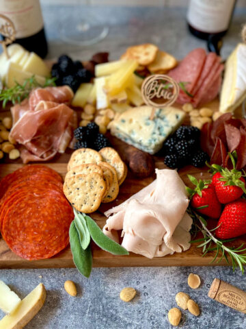 There is a large wooden board with a variety of food items: Meats, cheeses, strawberries, blackberries, nuts, olives, crackers, chocolate truffles, and fresh green herbs. There are four bottles of wine and three clear wine glasses. Items are on a light grey surface with a white time black wall.