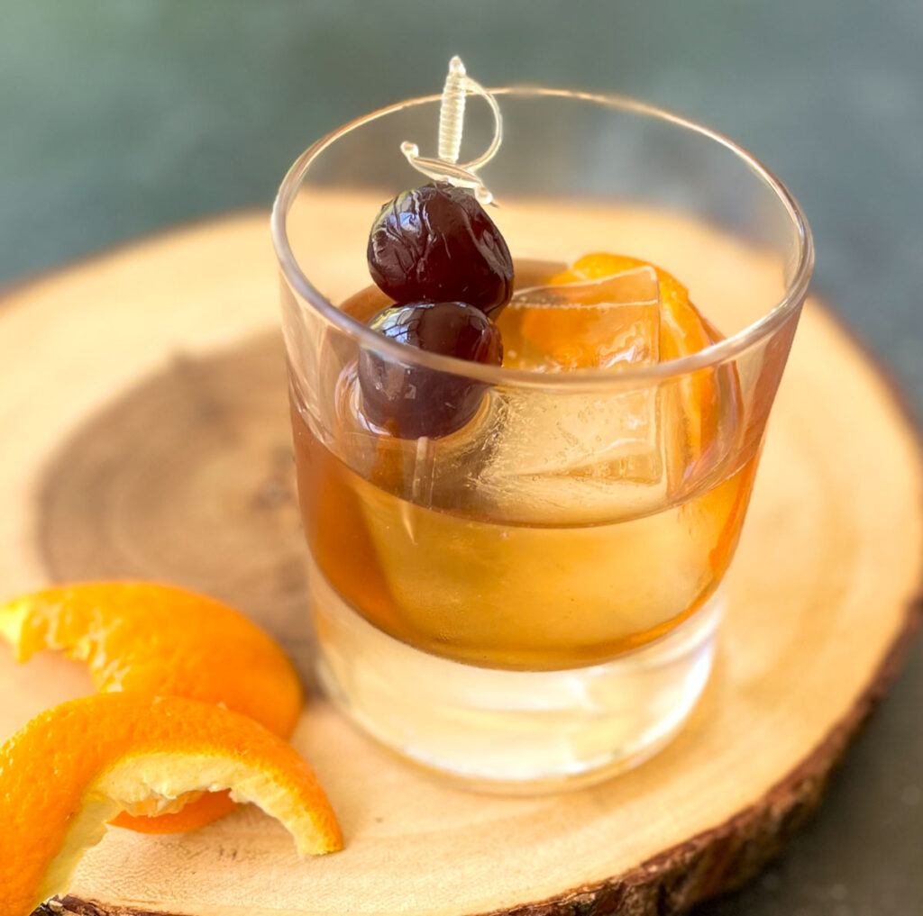 There is a short clear glass with light brown liquor, a large ice cube, an orange peel, and a cocktail pick with 2 dark red cherries on it it. There are 2 orange peels laying next to the glass. Items are on a round piece of wood. The piece of wood is on a gray surface.