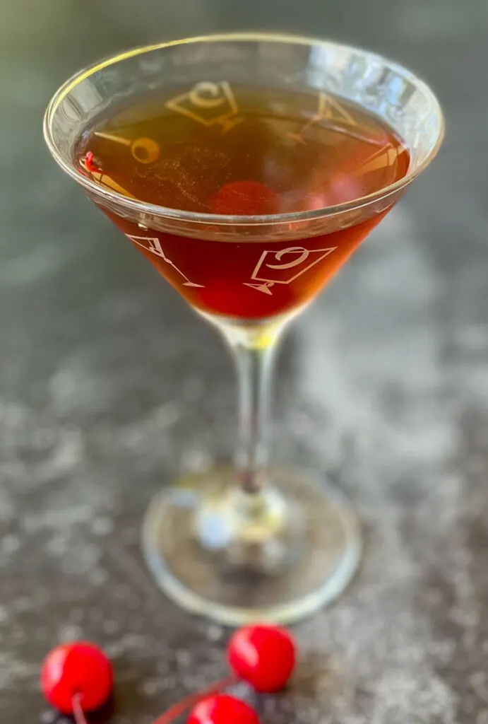 There is a stemmed cocktail glass with reddish brown liquor and a red cherry in it. There are three stemmed cherries laying in front of the glass. Items are on a dark gray surface.