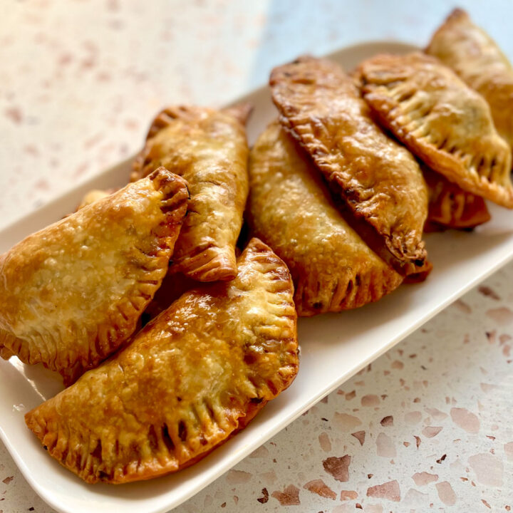 There is a white rectangular plate with 8 baked beef empanadas on it. The plate is on a light red and white terrazzo surface.