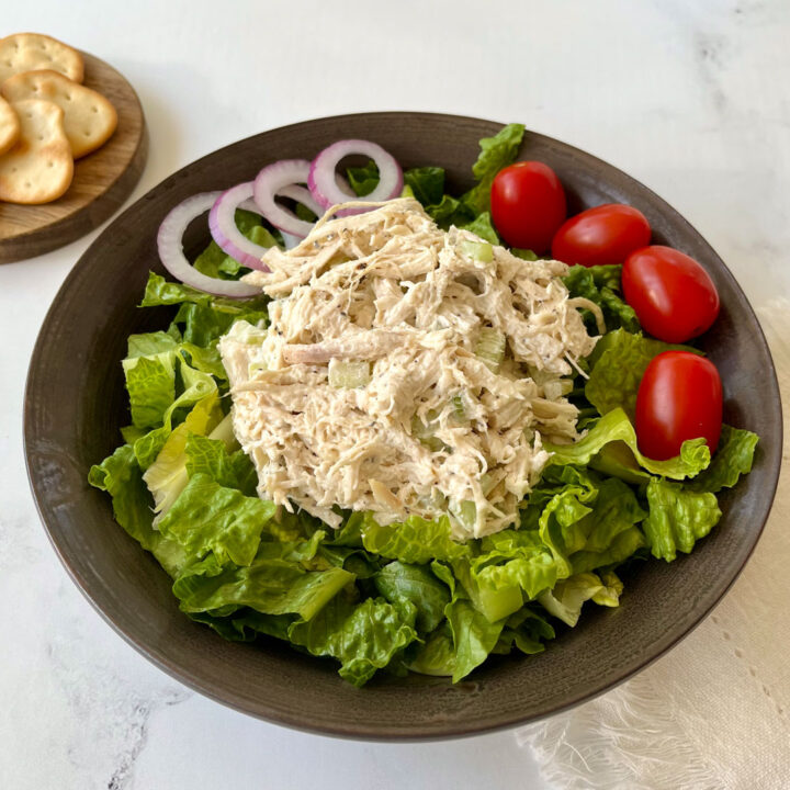 There is a brown bowl with chopped up lettuce in it. There is a large scoop of chicken salad on top of the lettuce. There are rings of red onion and four cherry tomatoes on the lettuce. There is a small round wooden board with light brown crackers on it in the background. Items are on a white marble surface.
