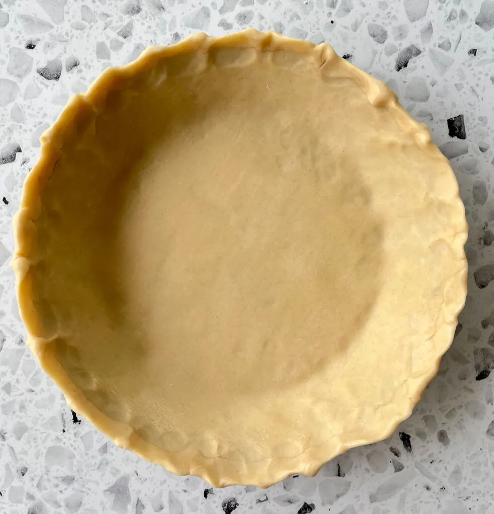 There is an unbaked pie shell in a pie pan. Pan is on a white and black terrazzo surface.