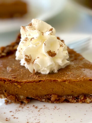 There is a small white plate with a piece of pumpkin pie with a ginger cookie and pretzel crumb crust on it. There is a dollop of white whipped cream and sprinkled cinnamon on top of the piece of pie. There is a stainless steel fork resting on the back of the plate. The plate is on a white marbled surface.