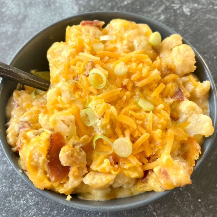 There is a gray bowl with pieces of cauliflower, melted cheese, pieces of bacon mixed together. There is shredded cheese and slices of green onion on top. There is a dark spoon handle resting inside the bowl . The bowl is on a dark gray surface.