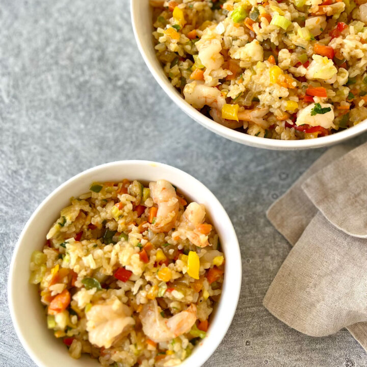 There is a white bowl with a combination of rice, shrimp, chopped up onion, garlic, red, yellow, orange, and green peppers. There is a larger white bowl in the background with the same ingredients in it. There is a light brown cloth napkin laying next to the bowl. Items are on a light gray surface.