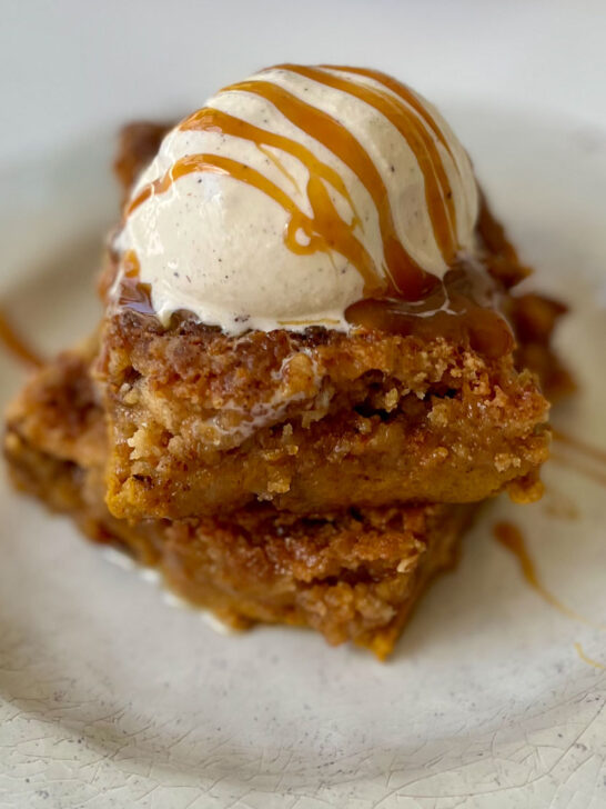 There is a tan plate with two squares of light brown cake with a scoop of vanilla ice cream on top and caramel sauce is drizzled over it. The plate is on a white marbled surface.