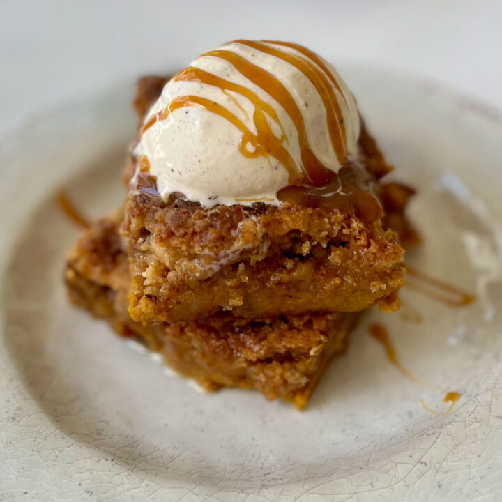 There is a tan plate with two squares of light brown cake with a scoop of vanilla ice cream on top and caramel sauce is drizzled over it. The plate is on a white marbled surface.