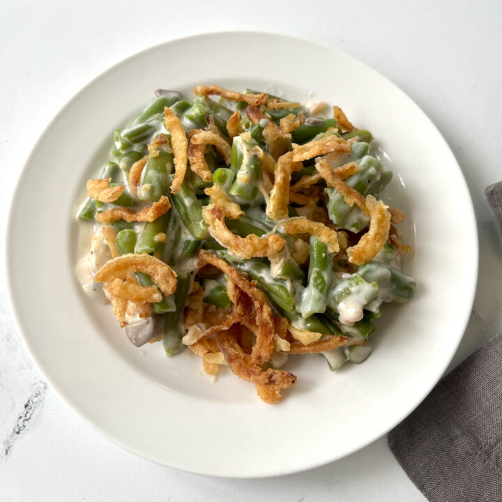 There is a small white plate with a scoop of a creamy green bean mixture with golden brown fried onion rings on top. There is a part of a gray cloth napkin laying to the right of the plate. Items are on a white marbled surface.