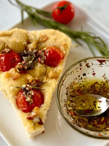 There is a square white plate with a pie shaped slice of thick bread. There are 3 cherry tomatoes, rosemary leaves, chunks of roasted garlic, and flakes of salt on top of the bread. There is also drizzled oil with herbs on top of the bread. There is a small clear glass bowl with oil and herbs in it. There is a small silver spoon resting in the bowl. There is a sprig of rosemary and a cherry tomato on the plate behind the bread. The plate is on a white marbled surface.