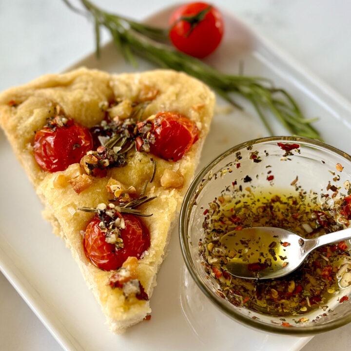 There is a square white plate with a pie shaped slice of thick bread. There are 3 cherry tomatoes, rosemary leaves, chunks of roasted garlic, and flakes of salt on top of the bread. There is also drizzled oil with herbs on top of the bread. There is a small clear glass bowl with oil and herbs in it. There is a small silver spoon resting in the bowl. There is a sprig of rosemary and a cherry tomato on the plate behind the bread. The plate is on a white marbled surface.