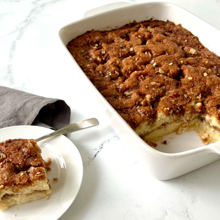 There is a white 9 inch by 13 inch baking dish with a yellow cake in it. There is a cinnamon, sugar, and chopped pecans topping on the cake. There is a square piece cut out of the cake. There is a small white plate next to the pan. There is a square piece of the coffee cake on the plate. There is a silver fork resting on the plate behind the piece of cake. There is a gray cloth napkin behind the plate. Items are on a white marbled surface.