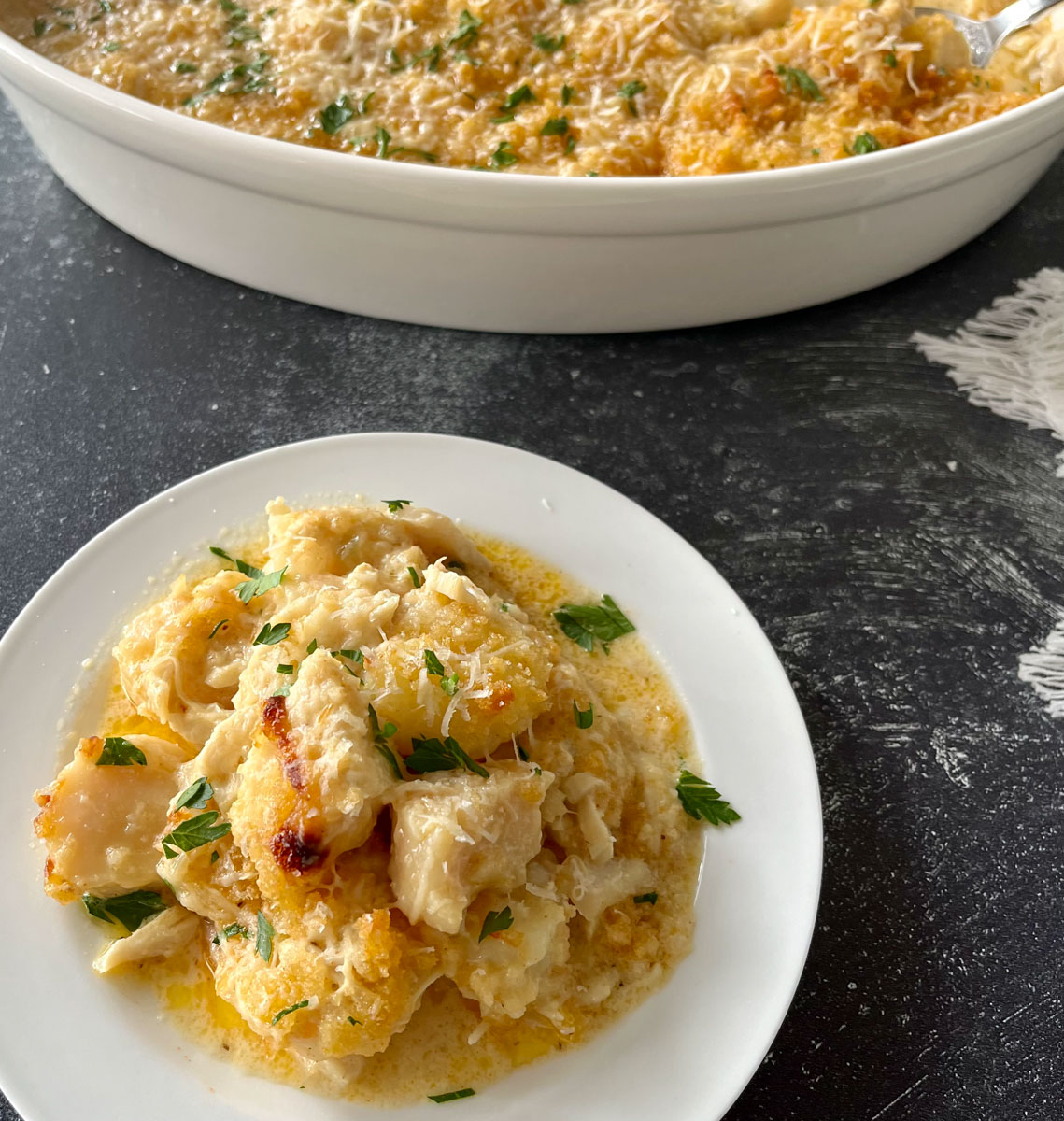 White round plate with baked pieces of white fish, sea scallops, shrimp, and crab meat in a buttery cream sauce. There is grated white cheese and chopped parsley leaves sprinkled over the top. There is a white oval casserole dish with the same seafood mixture in it. Items are on a gray surface.