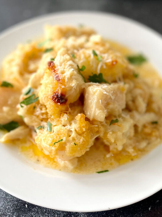 White round plate with baked pieces of white fish, sea scallops, shrimp, and crab meat in a buttery cream sauce. There is grated white cheese and chopped parsley leaves sprinkled over the top. Plate is on a gray surface.