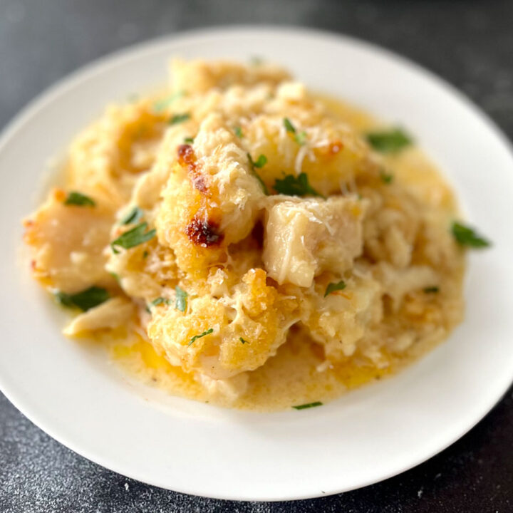 White round plate with baked pieces of white fish, sea scallops, shrimp, and crab meat in a buttery cream sauce. There is grated white cheese and chopped parsley leaves sprinkled over the top. Plate is on a gray surface.