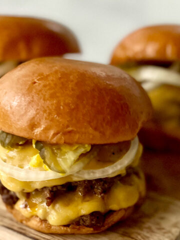 There are 3 hamburgers on a wood surface. Each burger has two thin beef patties, two slices of yellow cheese, onion slices, pickle slices, and mustard in between a bun.