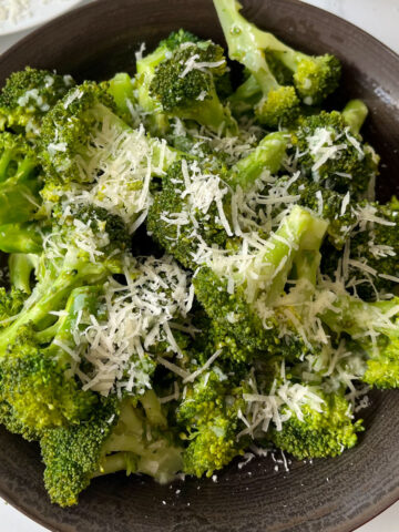 There is a large brown bowl with cooked broccoli florets in it . There is a light yellow sauce with minced garlic drizzled over the broccoli. There is also finely grated white cheese on top of the broccoli. There is a light tan cloth napkin next to the bowl. Items are on a white marbled surface.