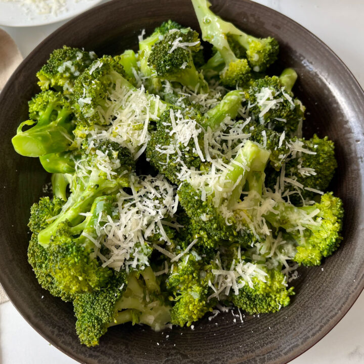 There is a large brown bowl with cooked broccoli florets in it . There is a light yellow sauce with minced garlic drizzled over the broccoli. There is also finely grated white cheese on top of the broccoli. There is a light tan cloth napkin next to the bowl. Items are on a white marbled surface.