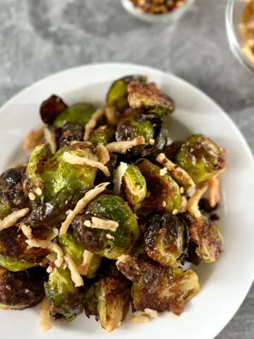 White plate with roasted golden brown small green cabbage looking Brussels sprouts that are cut in half. There is a dark brown sauce drizzled over the sprouts and pieces of fried onions on top. There is a small clear glass bowl with pieces of fried onions in it. Bowls are on a light gray surface.