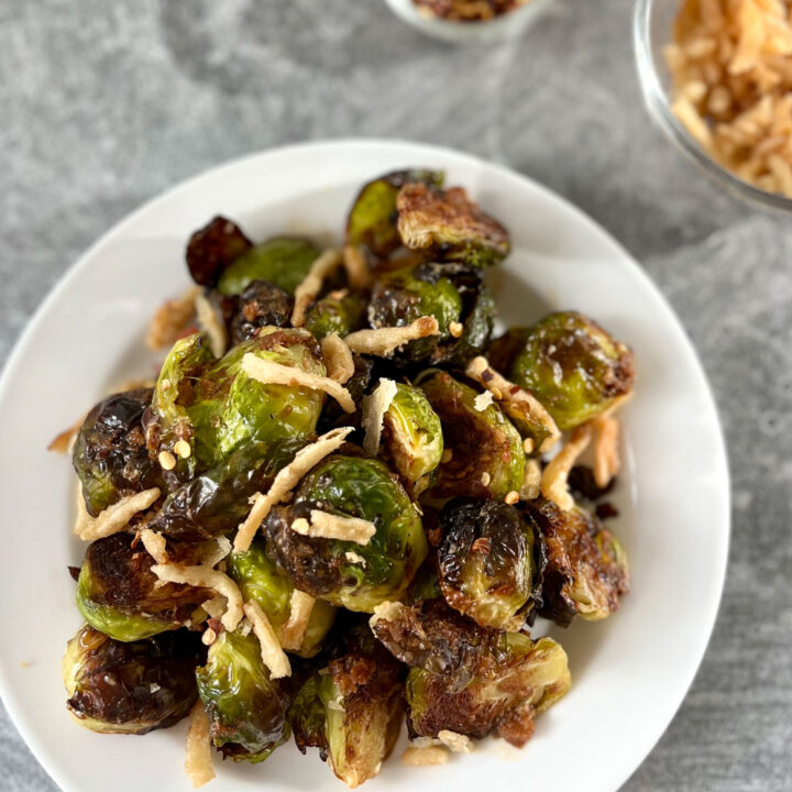 White plate with roasted golden brown small green cabbage looking Brussels sprouts that are cut in half. There is a dark brown sauce drizzled over the sprouts and pieces of fried onions on top. There is a small clear glass bowl with pieces of fried onions in it. Bowls are on a light gray surface.