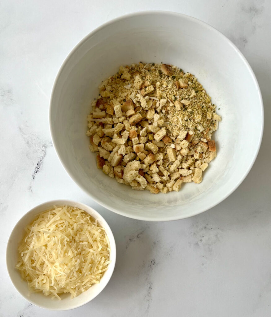 There is a large white bowl with pieces of seasoned dry bread. There is a medium size white bowl next to the large bowl with shredded white cheese in it. Bowls are on a white marbled surface.