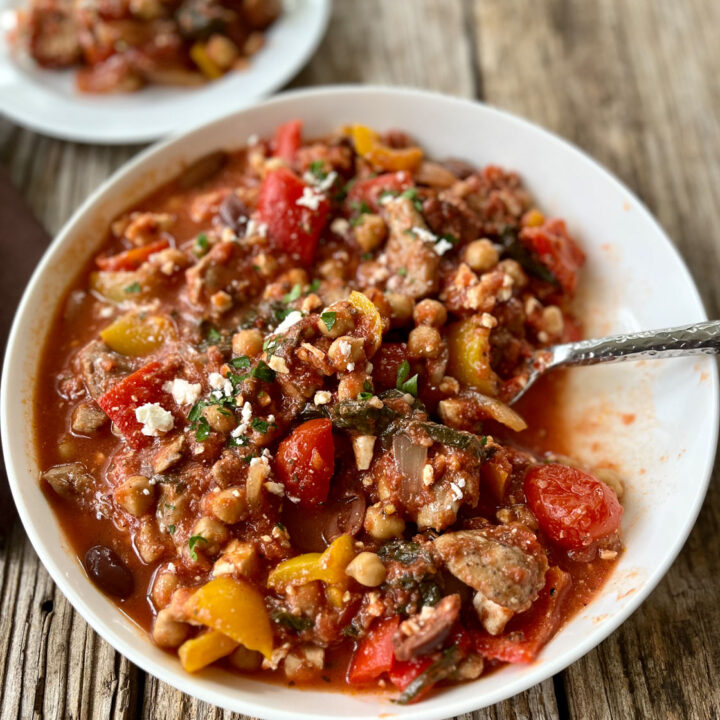Large white bowl with a variety of bell peppers, tomato sauce, pieces of chicken, spinach, white beans, and crumbled white cheese. There is a spoon resting in the bowl. There is a small white plate with the same mixture on it. Items are on a wood surface.