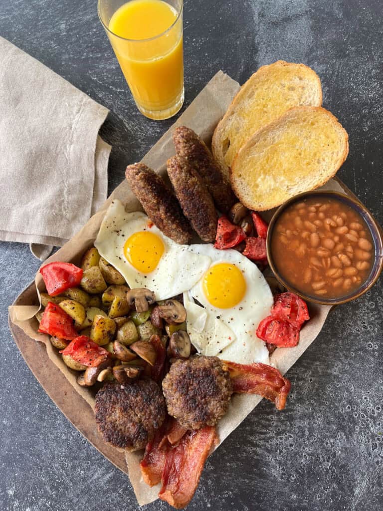 There is a rectangular tray lined with light brown parchment paper. There is a bowl of beans in brown sauce on the tray. There is 2 pieces of white toast with butter on them. There is 2 sausage patties, 2 fried eggs, cut up roasted tomatoes, sliced mushrooms, cut cup potatoes, sliced mushrooms, cut up roasted tomatoes, and three sausage links laying on top of slices of bacon. There is a tall clear glass with orange juice in it and a light brown cloth napkin. Items are on a dark gray surface.